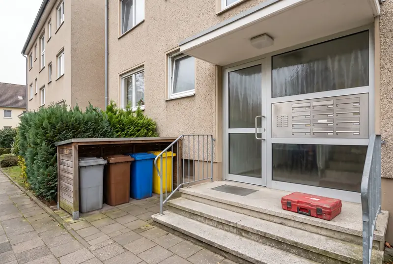 Hauseingang eines Mehrfamilienhauses mit Briefkastenanlage, gepflegten Stufen, Mülltonnenbox und abgestelltem Werkzeugkoffer
